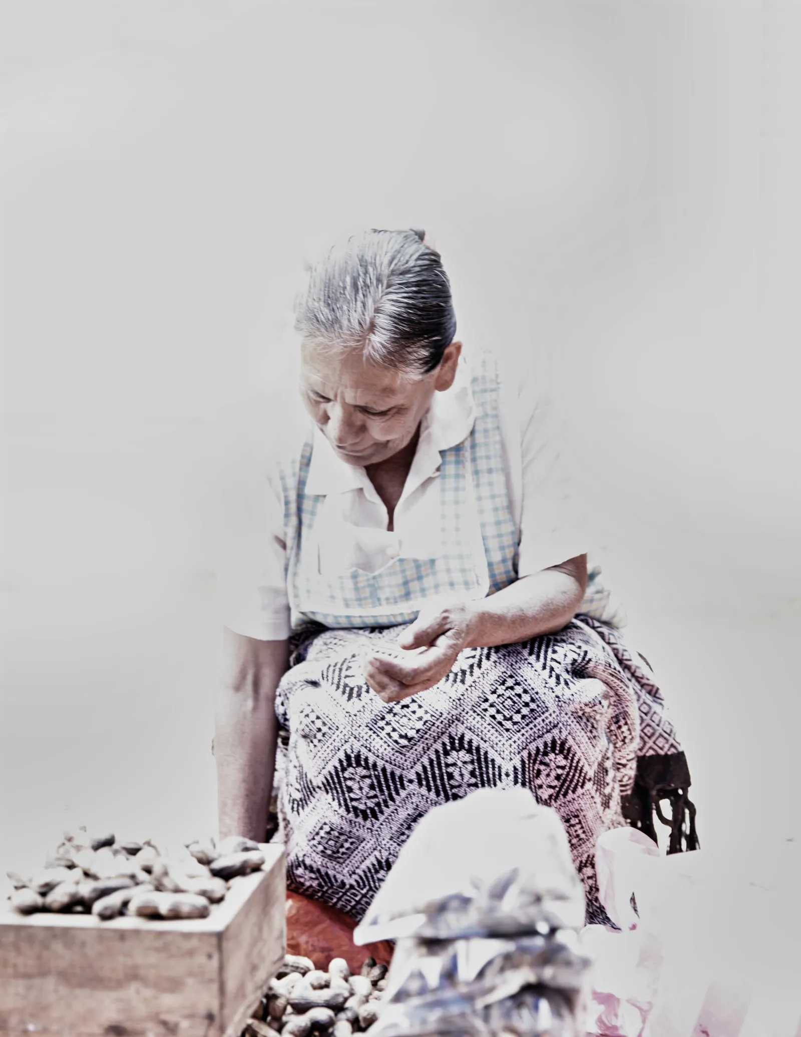 an older woman with gray hair looking down at her hands, seated beside a crate of nuts, in a washed-out pale light