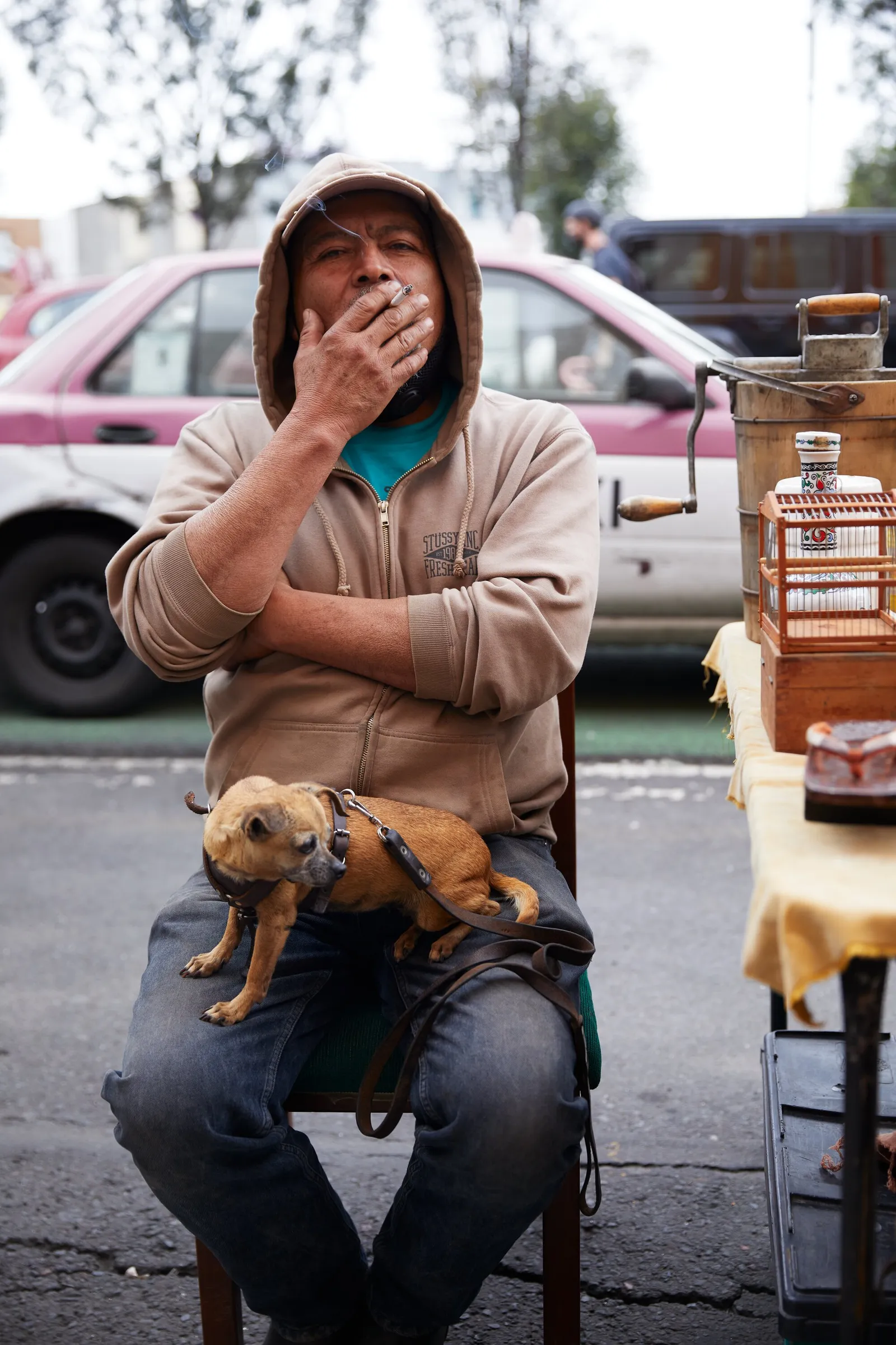 a man in a hooded sweatshirt seated on a chair holding a cigarette to his mouth, with a small tan dog on his lap and a parked car behind