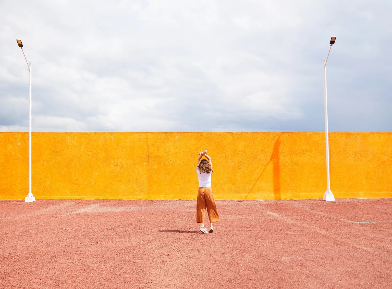 woman in white top and orange trousers stretching her arms overhead before a tall saturated yellow wall, with two white lamp posts on pink gravel
