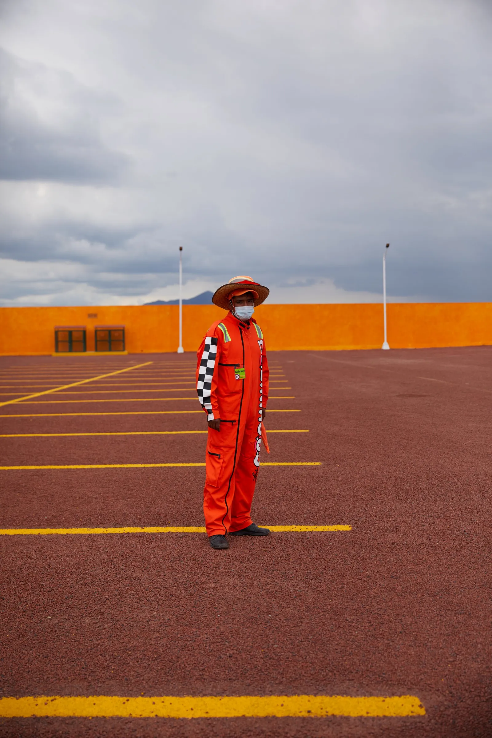 a person in an orange racing-style jumpsuit and face mask standing alone in an empty parking lot with yellow lines, orange wall and stormy sky