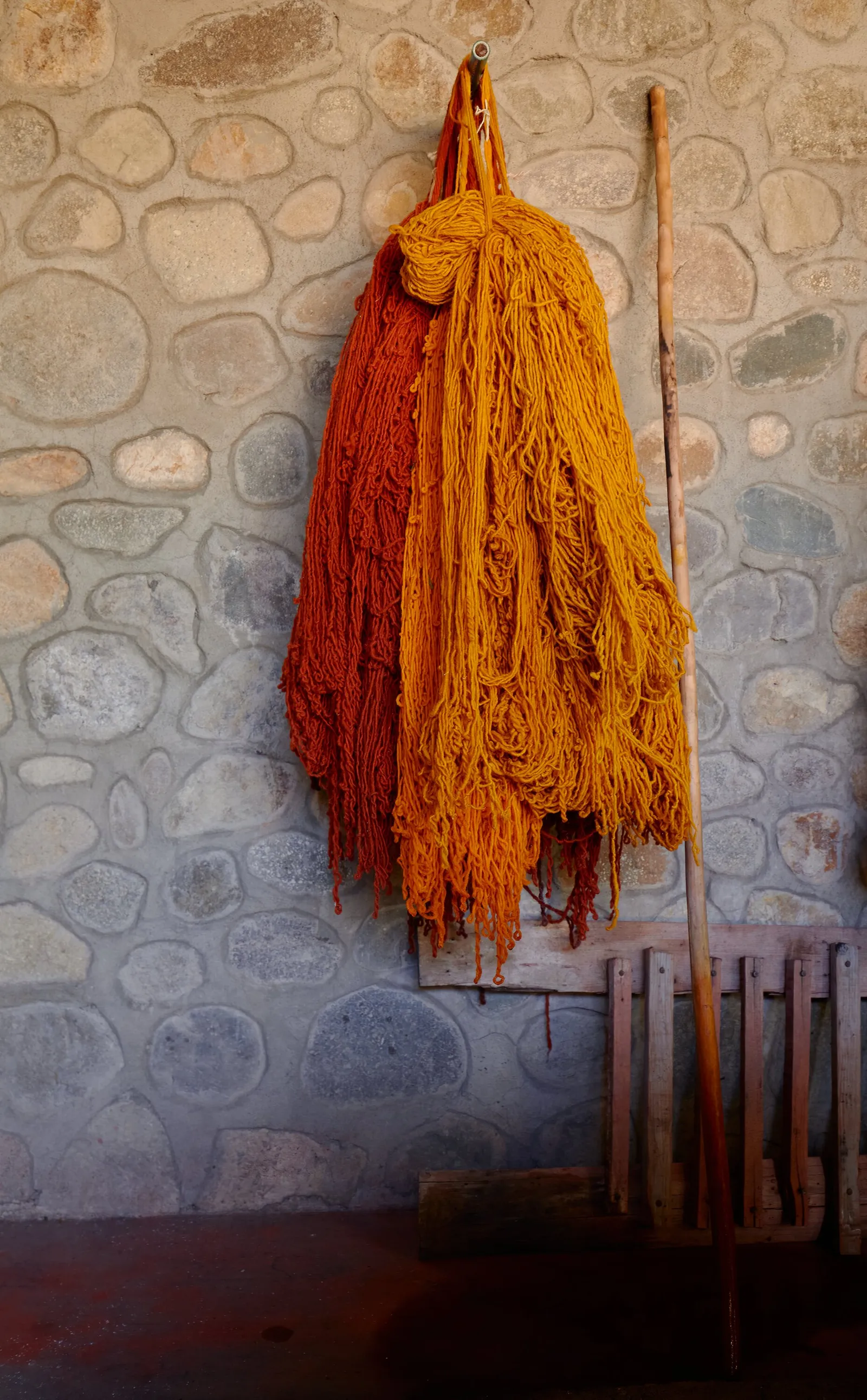 orange and rust-colored hanks of dyed wool hanging from a hook against a rough stone wall, with a wooden staff beside them
