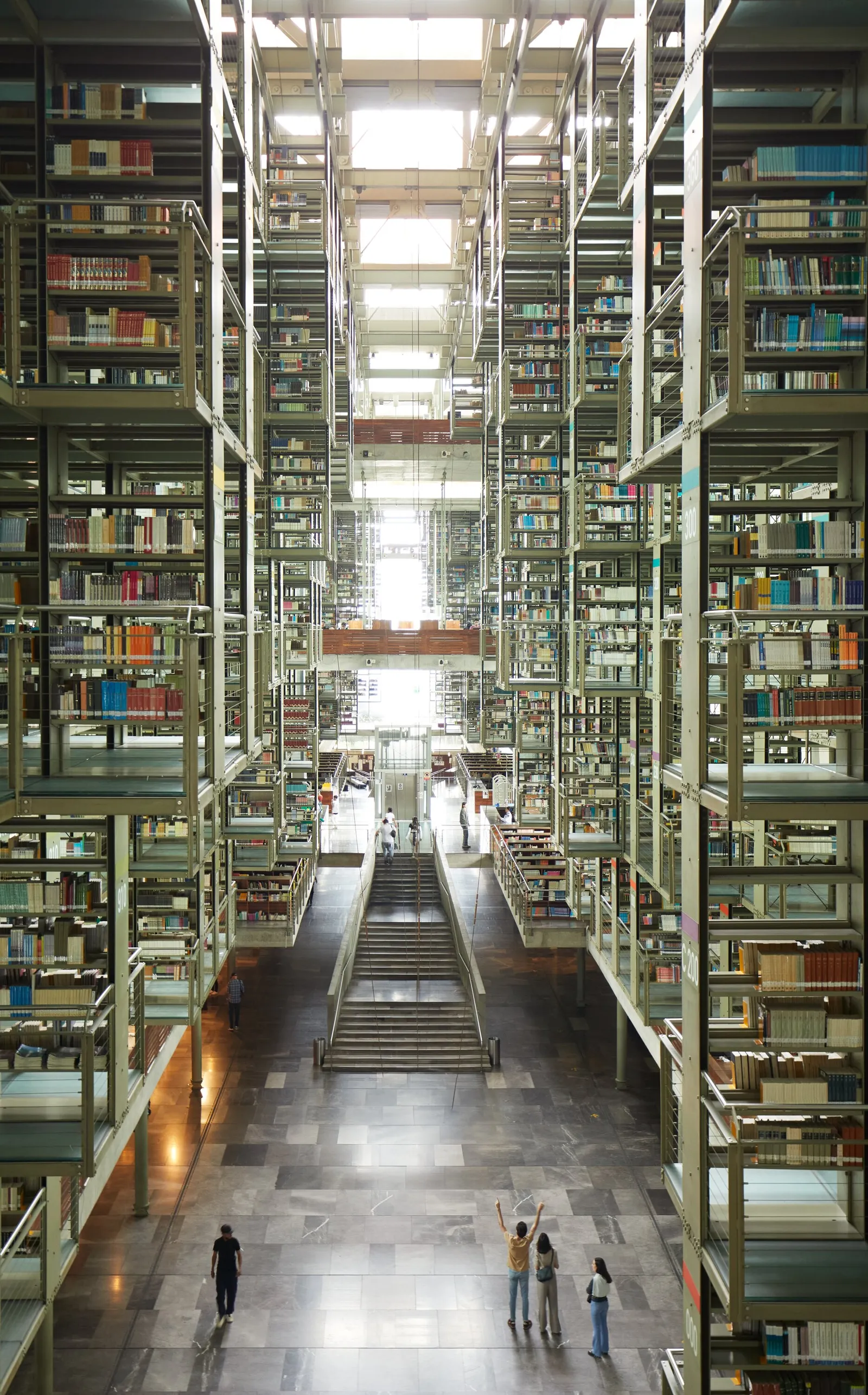vast library atrium with open metal book stacks rising on both sides, a central stair, and three people standing on the stone floor below