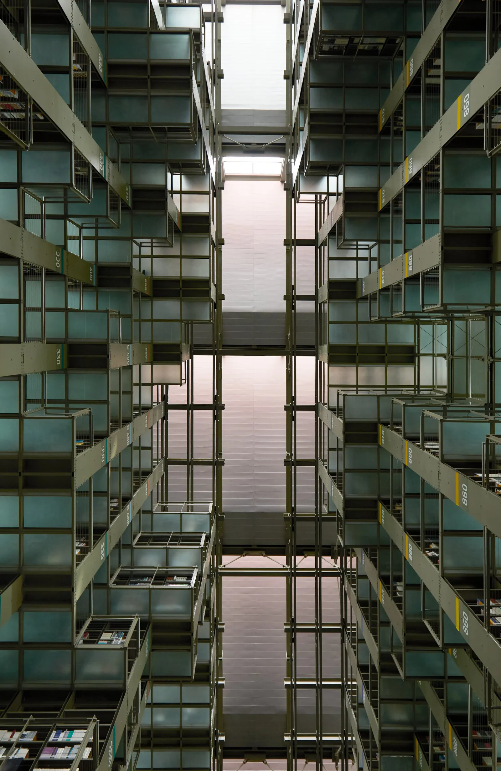 looking straight up between towers of green-tinted shelving and walkways toward a pale pink skylit ceiling