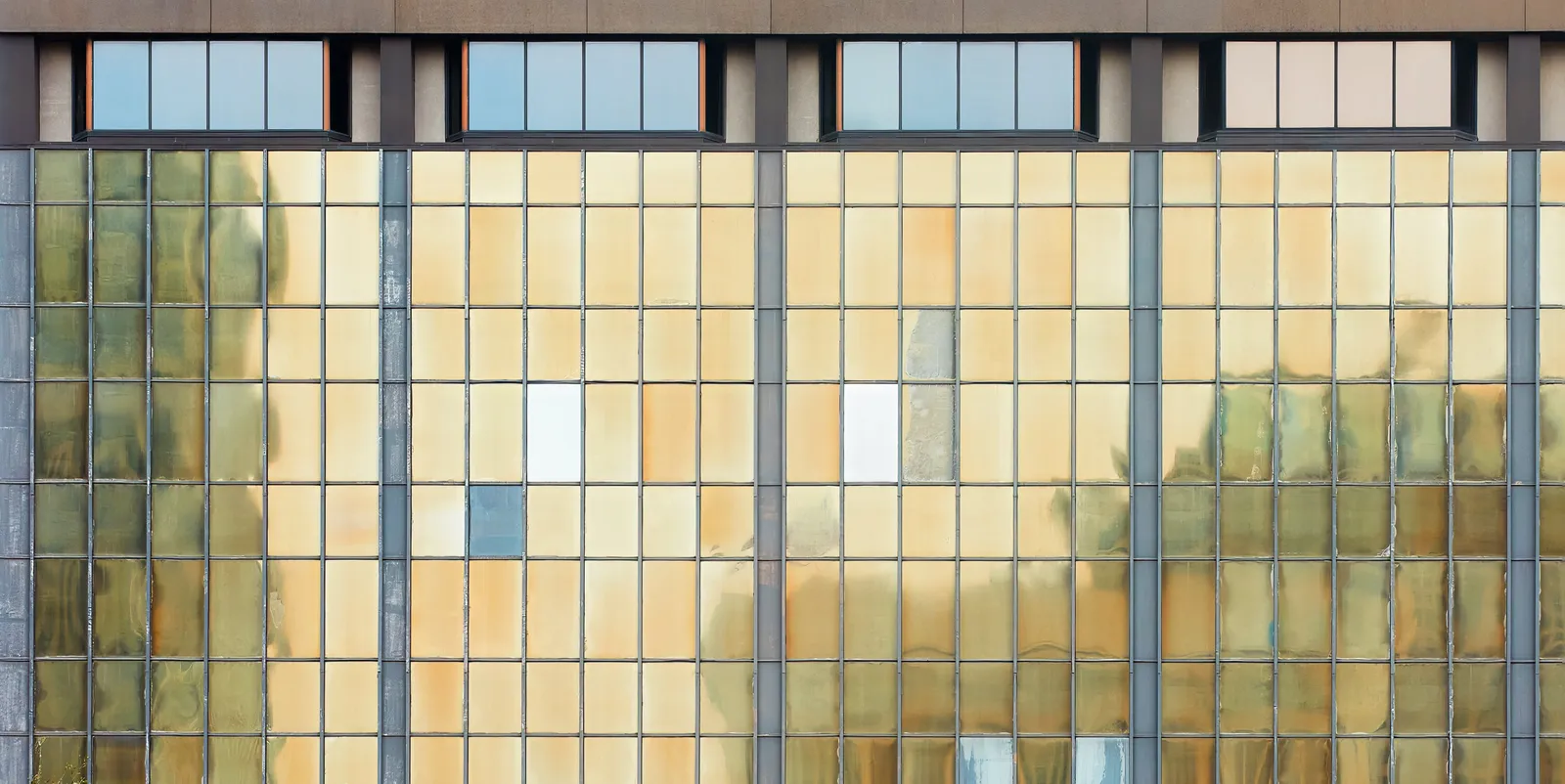flat glass facade of amber, green and cream panes in a grid of dark steel mullions, a few white and blue panes breaking the pattern