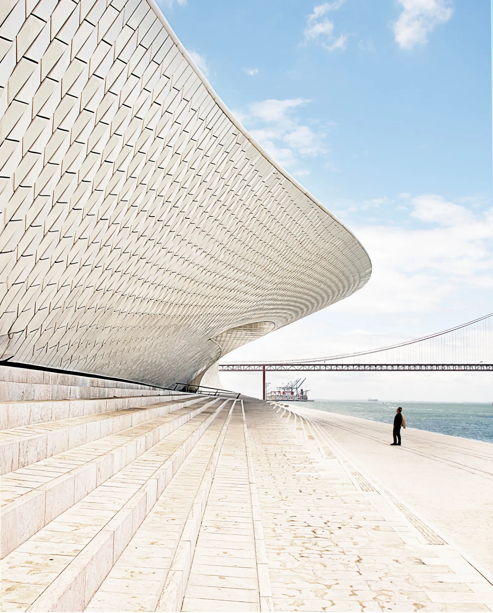sweeping white tiled building curving over wide stone steps, a lone figure on the waterfront promenade with a red suspension bridge beyond