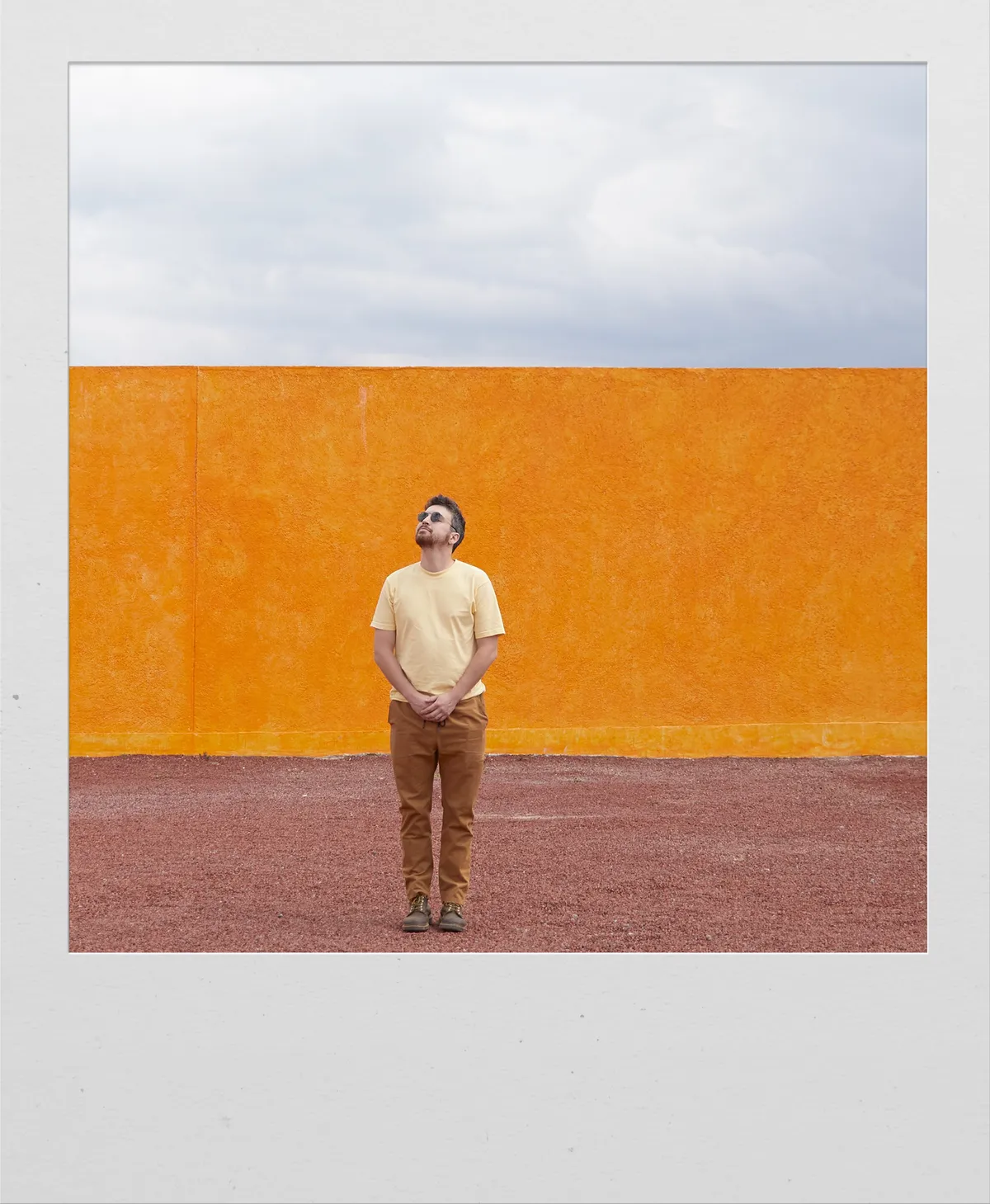 Polaroid of Casey Zane standing small in front of a tall orange wall, looking up at a cloudy sky